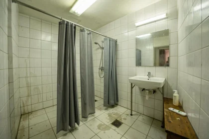 Salle de douches commune dans une auberge de jeunesse avec cabines séparées par des rideaux gris et carrelage blanc usé